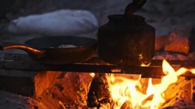 Kırsal bir açık hava yemek sahnesi, kararmış bir çaydanlık ve tava kükreyen bir kamp ateşinin üzerinde oturup sıcak ve davetkar bir atmosfer yaratıyor.