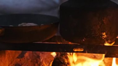 Tavuklu bir tava ve bir çaydanlık kamp ateşinin üzerinde kaynıyor, güneş batarken açık hava yemeklerinin sıcaklığını ve atmosferini yakalıyor..