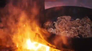 Tavuk parçaları cızırdar ve alevler tavayı yalayınca alevler yükselen bir kamp ateşi, duman ve buhar üzerinde dökme demir bir tavada pişirir. Kırsal açık hava aşçılık sahnesi.