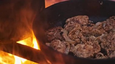 Cızırdayan tavuk ve soğanlı bir tava kamp ateşinin üzerinde pişiyor, dışarıda yemek pişirmenin kırsal çekiciliğini vurguluyor..