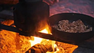 Kamp ateşinin üzerinde tavada kızaran tavuk ve soğanlara yakın. Alevler yanıp sönüyor ve büyüleyici bir görüntü yaratıyor..