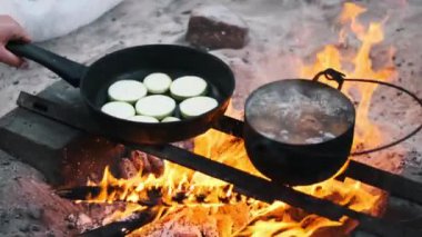 Bir el, kamp ateşinin üzerinden kızarmış kabak dilimleriyle kızartma tavasını kaldırıyor, açık hava yemekleri ve yemek hazırlıyor..