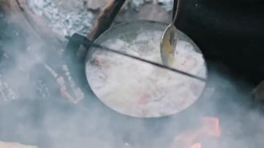 Adamın biri, kamp ateşinin üstünde tencerede çorba karıştırıyor. Basit açık hava yemekleri..