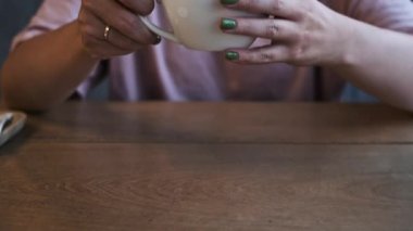 Bir kadın nazikçe bir bardak kapuçinoyu ahşap bir masaya koyar, kremalı köpükle hala görülebilen narin latte sanatı. Sahne bir sakinlik ve rahatlama hissi uyandırıyor..