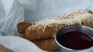 Cömertçe parmesanla kaplanmış çıtır peynirli çubukların yakın plan çekimi, canlı kırmızı bir sos eşliğinde, izleyiciyi dokuları ve lezzetleriyle cezbediyor..