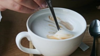 Bir kadının, beyaz bir fincanda kremalı latte kahvesini nazikçe karıştırırken, kıvrımlı köpük desenlerini ortaya çıkarırken, üst görüntü yavaş çekim.