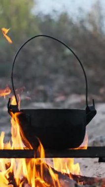 Alevli bir kamp ateşinin üzerinde asılı duran siyah metal bir tencerenin dikey videosu. Alevler tencerenin dibini yalayarak sıcak ve davetkar bir atmosfer yaratıyor..