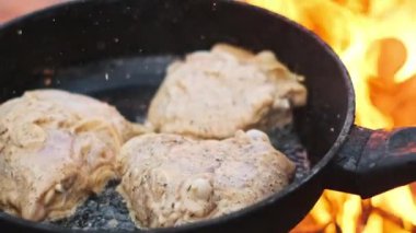 Bir tavada cızırdayan tavuk kalçalarının yakın çekim, alevler etrafında yükselirken, açık havada pişirmenin özünü yakalıyor..