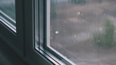 Pencereye düşen yağmurun durağan bir kamerayla çekilmiş sakin görüntüleri. Bulanık arka plan yağmurlu bir manzarayı ortaya çıkarıyor, barışçıl ve düşünceli bir atmosfer yaratıyor..