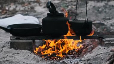 Kamp ateşi, siyah bir çaydanlık ve tencereyle birlikte, dışarıda pişirilmeye hazır metal bir ızgaranın üzerinde parlak bir şekilde yanar..