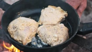 Üç parça terbiye edilmiş tavuk bir kamp ateşinin üzerinde tavada cızırdıyor. Kırsal açık hava yemekleri..