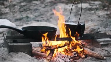 Bir tava ve bir tencere çatırdayan bir kamp ateşinin üzerinde oturur, alevler altında dans eder, rahat ve davetkar bir yemek sahnesi yaratır..