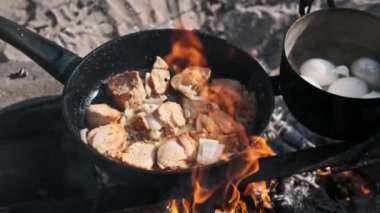 Bir tencere cızırdayan et ve soğan bir kamp ateşinin üzerinde pişiyor. Kaynayan bir yumurta tenceresinin yanında, kırsal bir açık hava yemek deneyimi yaratıyor..