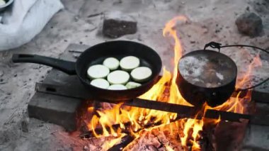 Bir tavada dilimlenmiş kabak ve bir tencere kaynar su kamp ateşinin üzerinde kaynayarak basit ve kırsal bir yemek pişirme sahnesi yaratıyor..