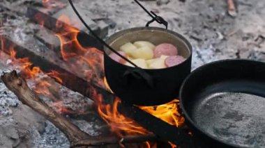 Patates ve sosisle dolu bir tencerenin yakın çekimi çıtırdayan bir kamp ateşinin üzerinde kaynayan, basit ve sağlıklı açık hava yemeklerinin özünü yakalıyor..