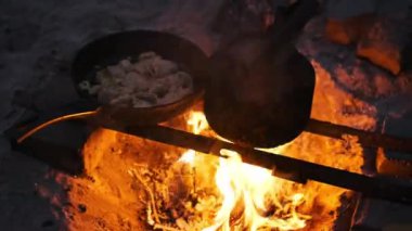 Yıldızlı bir gece gökyüzünün altında, kırsal bir kamp ateşi kararmış bir çaydanlığı ve cızırdayan bir tavuğu aydınlatır, açık havada pişirmenin ve maceranın basit zevklerini gözler önüne serer.