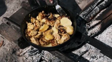 Bir kadının eli, tavukla patatesleri karıştırır. Kamp ateşinin üstünde demir bir tavada kızartır..
