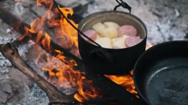 Patates güveci ve sosis çıtırdayan bir kamp ateşinin üzerinde kaynıyor ve açık hava yemeklerinin özünü yakalıyor..