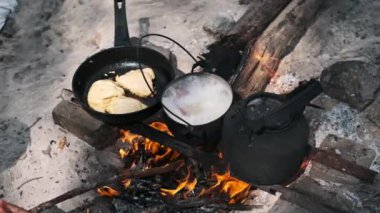 Tavada tavuk cızırtısı ve tencerede çorba köpükleri, ikisi de canlı bir kamp ateşinde pişiyor. Yakınlarda eski bir çaydanlık duruyor..
