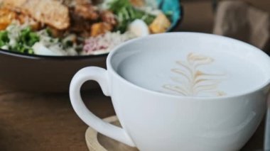 Bir kafede, ahşap bir masaya konmuş sütlü kapuçino ve latte sanatının yakın plan çekimi. Odak, izleyiciyi davetkar aroma ve aromayı hayal etmeye davet eder..