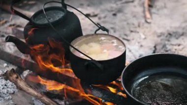 Sıcak bir tencere yahni, yağlı siyah bir tava ve kükreyen bir kamp ateşiyle kırsal bir açık hava yemek sahnesi..