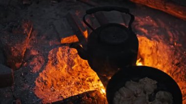Parlayan közlerden bir yatağın üzerinde yemek pişiren demir çaydanlık ve tava büyüleyici ve kırsal bir kamp ateşi sahnesi yaratıyor..