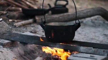 Siyah kazan bir kamp ateşinin üzerinde duruyor, içinde su kaynıyor. Hayatta kalma, doğada yemek pişirme, basit yaşam konsepti.