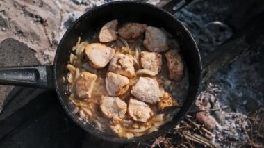 Tavuk ve soğan parçaları çatırdayan bir kamp ateşinin üzerinde kaynıyor. Sahnede kırsal ve geleneksel yemek pişirme stili sergileniyor.