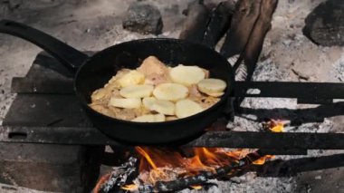 Dilimlenmiş patatesler, canlı bir kamp ateşinde pişen tavuk butlarına eklenir..