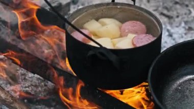 Patates dolu bir tencere ve sosis kükreyen bir kamp ateşinin üzerinde kaynıyor. Açık hava meraklıları için doyurucu bir yemek..