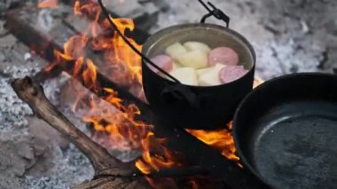 Bir tencere patates ve sosis canlı bir kamp ateşinin üzerinde kaynıyor ve doğanın kalbinde hazırlanmış klasik ve rahatlatıcı bir yemek sergileniyor..