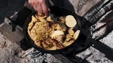 Tavuk ve patatesleri karıştırmak için çatal kullanan bir kadının el manzarası. Kamp ateşinin üzerinde kızgın demir tavada kızartılıyor..