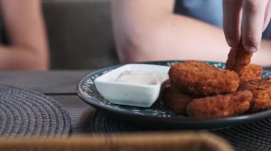 Bir çocuğun tavuk nuggetını alıp beyaz sosa batırırken yan görüntüsü. Külçeler mavi desenli bir tabağa yerleştirilmiş..
