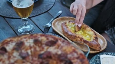 Bir kadının elinin Adjarian Khachapuri 'den bir parça koparıp arka planda bira ve pizzayla erimiş peynir ve yumurta sarısına batırdığı yakın plan bir fotoğraf..