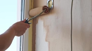 Bir kadının eli, pencerenin yanındaki bej boyalı beyaz bir duvarı boya rulosu kullanarak boyar. Onarım ve yenileme konsepti.