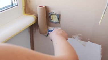 Bir kadın, maskeli bir elektrik prizinin etrafındaki hassasiyete odaklanmış bej rengi bir duvarı silindirle beyaza boyuyor. Ev yenileme ve DIY konsepti.