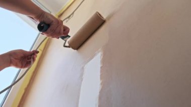 Düşük açıdan, bir boya silindiri bej bir duvara beyaz boya uygular. Sarı maskeleme bandı pencere çerçevesini korur. Bu çekim ev yenileme ve DIY projelerini somutlaştırıyor.