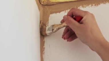 Bir kadın, bej boya sürerek bir duvar köşesini fırçayla dikkatlice boyuyor. Yenileme işleminin kapanışı.