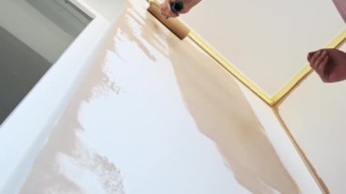 Bir kadın boya silindiri kullanarak duvar bejini boyuyor. Kenarları maskeleme bandıyla korunuyor, düzgün bir son garantiliyor. Düşük açı, yenileme süreci hakkında ilginç bir perspektif sağlıyor.