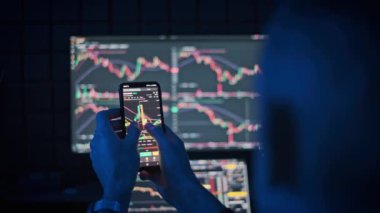 Bir kripto tüccarı gerçek zamanlı pazar verilerini izlemek için bir telefon kullanır ve arka planda bilgisayar ekranında ticaret grafikleri görüntülenir. Finans ve teknoloji projeleri için ideal.