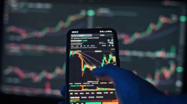 Bitcoin fiyat tablosunu gösteren bir cep telefonunun yakın çekimi. Arkaplan, bir bilgisayar ekranında pazar etkinliğini vurgulayan bulanık ticaret grafikleri içeriyor.