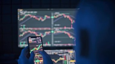 Bir kripto tüccarı, pazar eğilimlerini izlemek ve loş aydınlatılmış bir odada Bitcoin fiyat dalgalanmalarını analiz etmek için mobil bir uygulama kullanır.