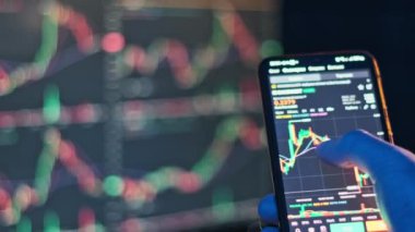 Bir tüccar, arka planında bulanık bir bilgisayar ekranı olan şifreli para şemasını izlemek için akıllı telefon kullanır. Fintech ve takas videoları için uygun.