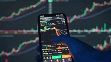 Cep telefonundaki Bitcoin fiyat grafiğini bilgisayar ekranındaki ticari grafiklerin bulanık arkaplanıyla karşılaştıran bir elin yakın plan görüntüsü.