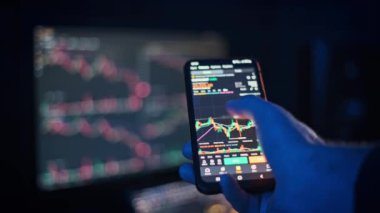 Arka planda bulanık bir bilgisayar ekranı olan şifreli para işlem çizelgelerini analiz etmek için akıllı telefon kullanan bir elin yakın çekimi. Fintech ve yatırımlarla ilgili projeler için mükemmel.