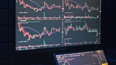 Kripto para çizelgeleri karanlık bir odadaki büyük bir bilgisayar monitöründe sergilenir. Benzer grafikleri gösteren bir dizüstü bilgisayar hafifçe görünür.