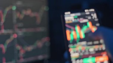 Bilgisayar ekranındaki borsa grafiklerinin bulanık arkaplanıyla şifreli para ticareti yapmak için cep telefonu kullanan bir elin yakın plan görüntüsü.