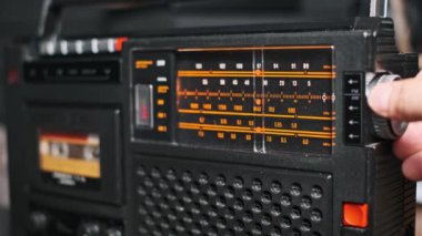 Antika bir radyoda analog kadranı bir kasetçalarla ayarlayan bir elin yakın çekimi. Nostaljik tasarım ve dokunsal detayları vurguluyor, retro, 80 'lerin nostaljisi veya klasik temalı projeler için ideal.