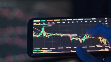 Bir parmak, akıllı telefon ekranındaki kripto para çizelgelerini pazar verilerinin bulanık arkaplanıyla karşılaştırır. İş ve finans içeriği için mükemmel.