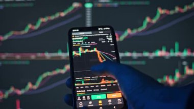 Bilgisayar ekranında pazar verileri gösterilen akıllı bir telefondaki Bitcoin çizelgelerinde gezinen bir elin yakın çekimi. Şifreleme ve takas videoları için mükemmel.